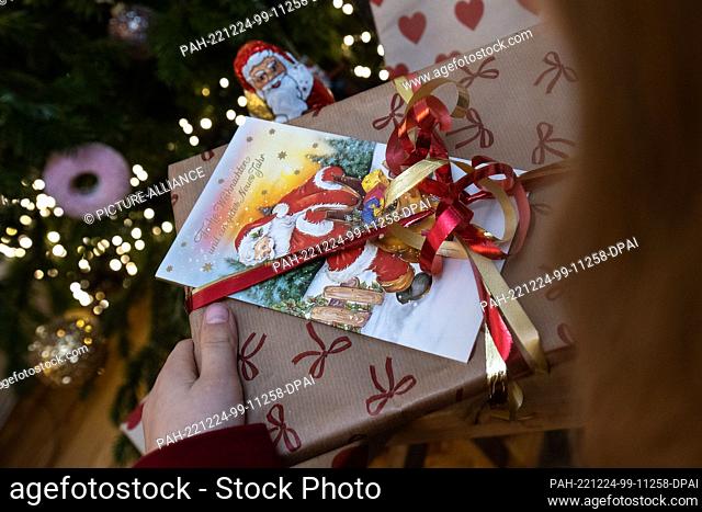 24 December 2022, Lower Saxony, Göttingen: A girl holds a gift in front of a decorated Christmas tree. In Germany, Christmas Eve has become the day for...