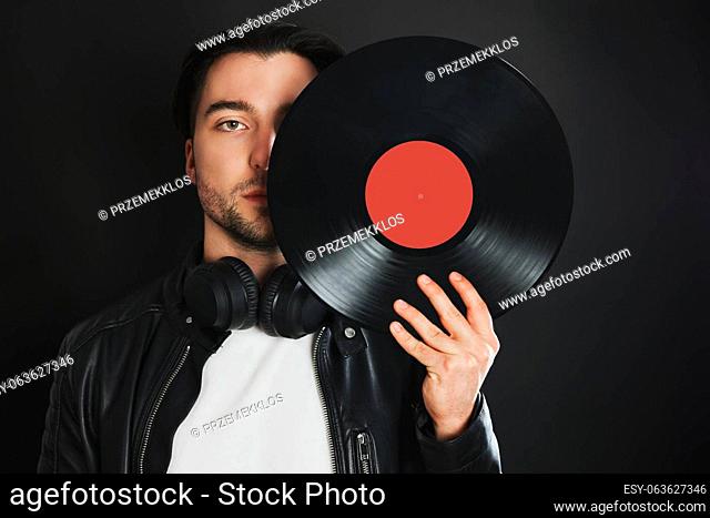 Man holding retro vinyl record covering face. Rock style. Vintage music style. Male wearing black jacket holding vinyl record disc standing on dark background