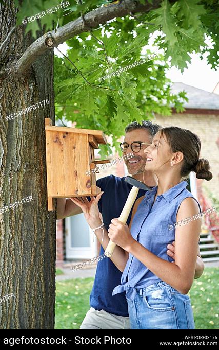 Happy girl with father hanging nest box on tree trunk in garden