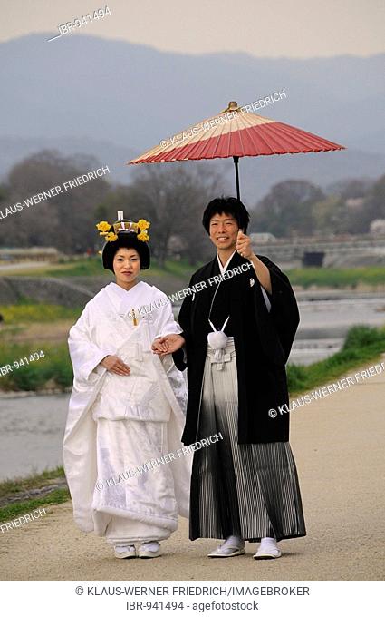 Traditional Japanese wedding couple wearing kimonos, bride with wedding hairstyle, groom holding a traditional paper parasol on the Kamigamo River, Kyoto, Japan