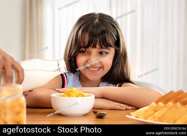A SMALL GIRL HAPPILY SITTING AND LOOKING AT MILK BEING SERVED