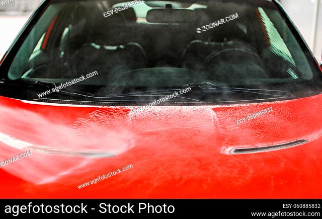 Front hood of red car washed in self service carwash, white water spraying on metal, blurred seats and interior background