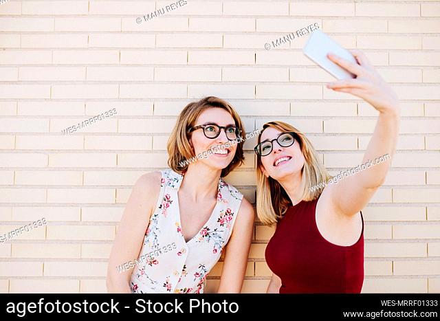 Smiling female colleagues taking selfie through smart phone in front of brick wall