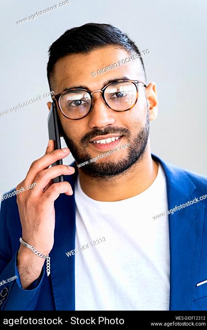 Businessman with eyeglasses talking on smart phone by wall in office