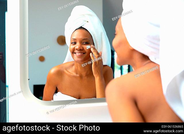 Smiling young african american woman applying face cream while looking in mirror, copy space