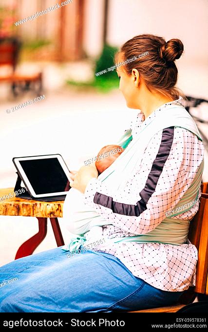 Young mother with little baby in sling sitting at cafe outdoors and working using tablet. Young multitasking mother