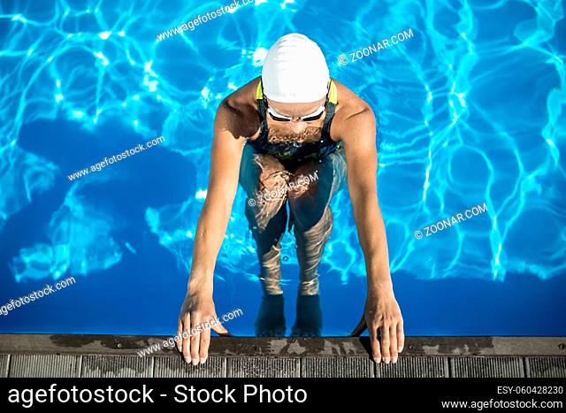 Sports girl in the swimming pool. Swimmer wears a black-lime swimsuit, a white swim cap and swim glasses. She looks forward and holds her hands on the pool side