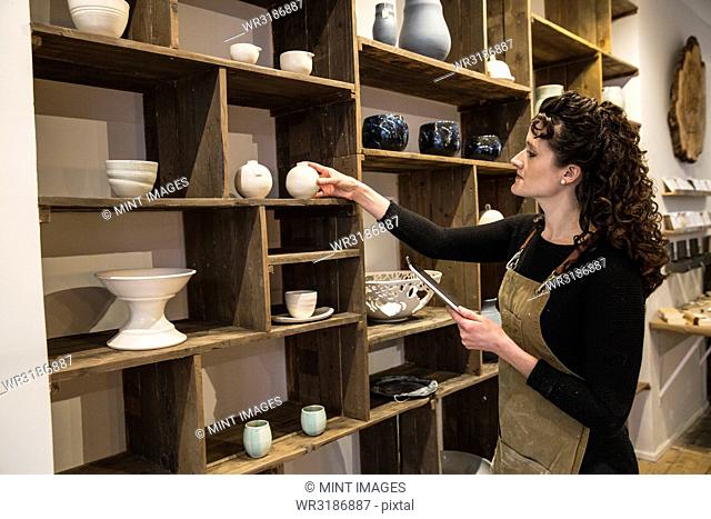 Woman with curly brown hair wearing apron standing in her pottery shop, arranging ceramic items on shelves, holding digital tablet