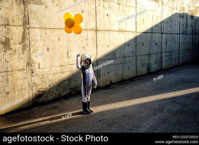 Little girl wearing space helmet holding balloon standing on street against wall