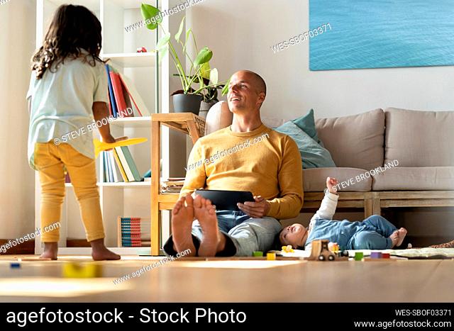 Father with digital tablet looking at toddler while baby lying by him at home