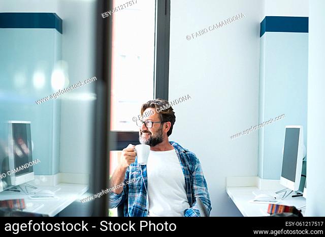 Adult man portrait sitting at desktop office doing job break drinking coffee and looking outside the window - online freelance employee lifestyle activity -...