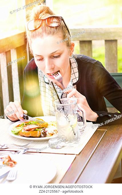 Sun flare image of an Australian person sitting on outdoor restaurant deck enjoying ham and salad sandwich with cutlery. Summer diner