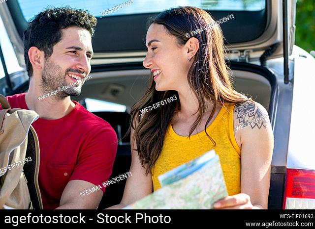 Smiling girlfriend looking at boyfriend while sitting in car trunk