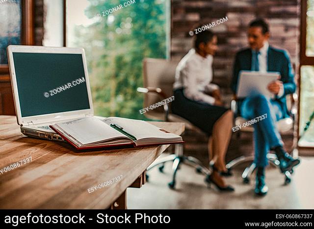 Laptop And Notebook With Notes Are On The Office Table, Couple Of Business People, Man And Woman Sitting On Blurred Back, Close Up, Toned Image