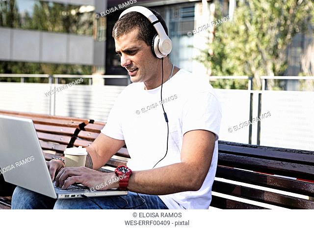 Young man sitting on a bench wearing headphones and using laptop