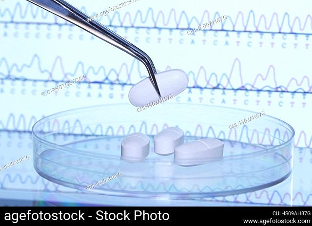 Tweezers holding pill over Petri dish. A computer screen that displays results of automated DNA sequencing is in the background