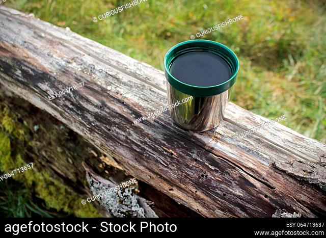 Green plastic thermos stainless steel cup put on a log on sunny day surounded by green grass in a forest