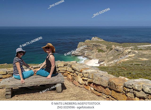 Two women sitting on a bench at cape of good hope enjoying the view over the ocean turnig and looking in to the camera, Cape of good hope, Cape Town