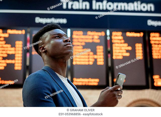 A businessman looking up in a train station while holding his mobile phone. The arrival departure board is behind him