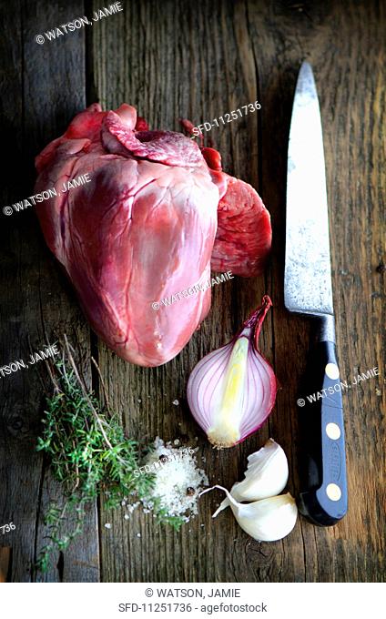 A pig's heart on a wooden board with an old knife, onions, garlic, thyme and salt