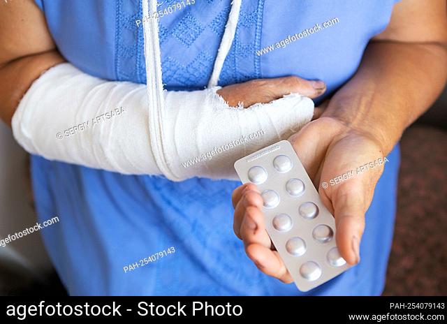 A senior retired woman with a rewound arm in a cast and a bandage holds anesthetic pills. Blow, fracture, bones, hospital || Model approval available