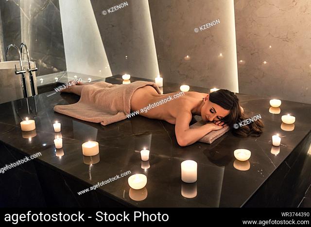 Beautiful woman lying down on massage table surrounded by scented candles in luxury spa and wellness center
