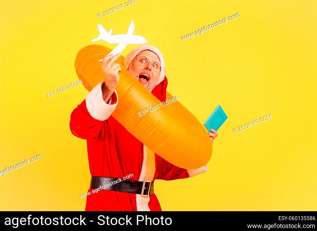 Portrait of amazed elderly man in santa claus costume standing with rubber ring, passport and paper plane, being happy to travel on Christmas holidays