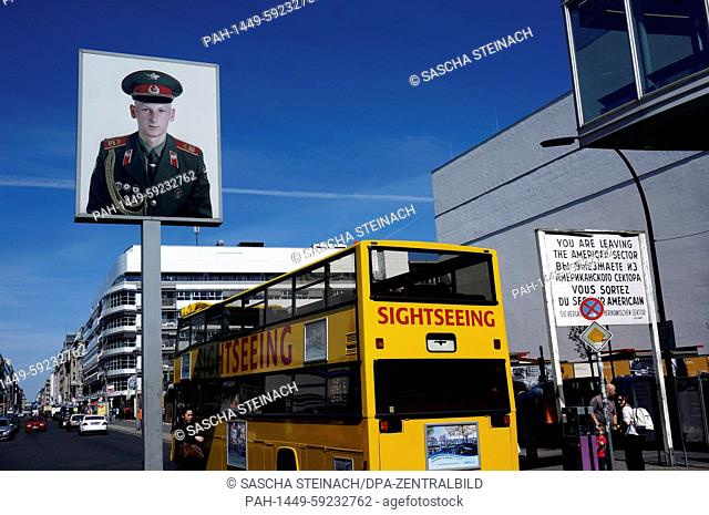 A yellow sightseeing bus passes a light box featuring a portrait of a Soviet soldier at Checkpoint Charlie on Friedrichstrasse in Berli