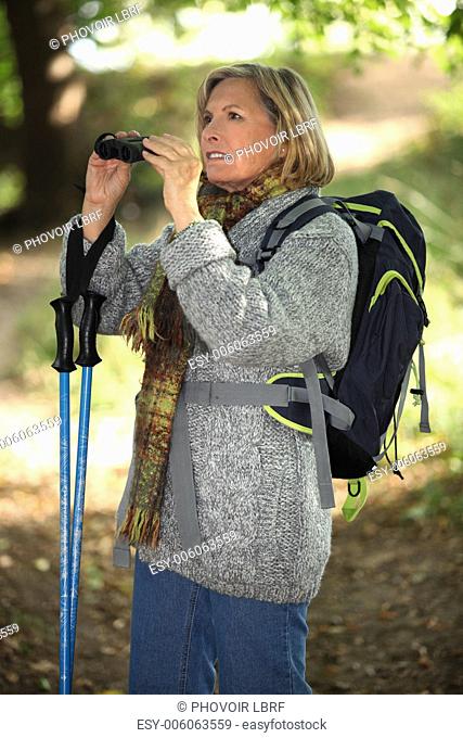 60 years old blonde woman is contemplating landscapes with binoculars, she's wearing warm clothes and using a crook