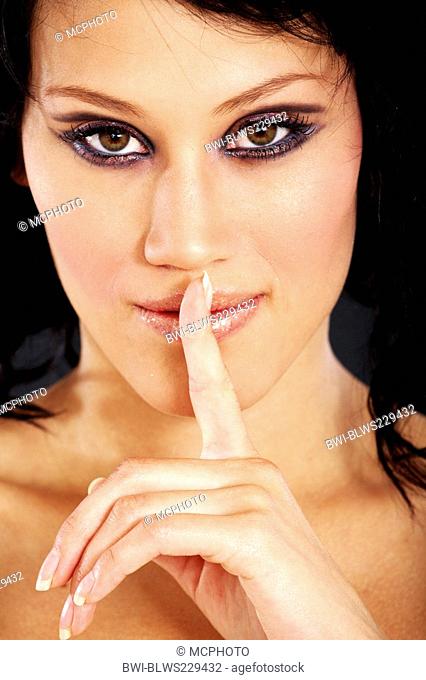young woman with eye-catching eye make-up laying a forefinger on her mouth, smirking