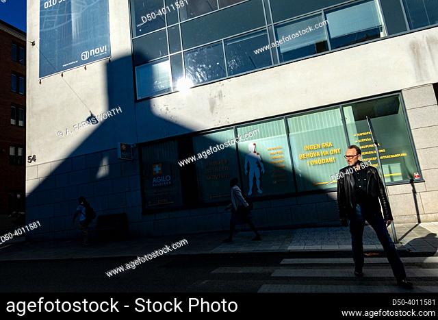 Stockholm, Sweden A man in a leather jacket crosses the street in strong sunlight casting a house shadow against a wall
