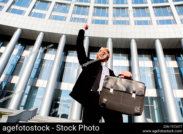 Close-up portrait of successful businessman with his arm up. Man in black suit posing with brief case isolated on office buiding