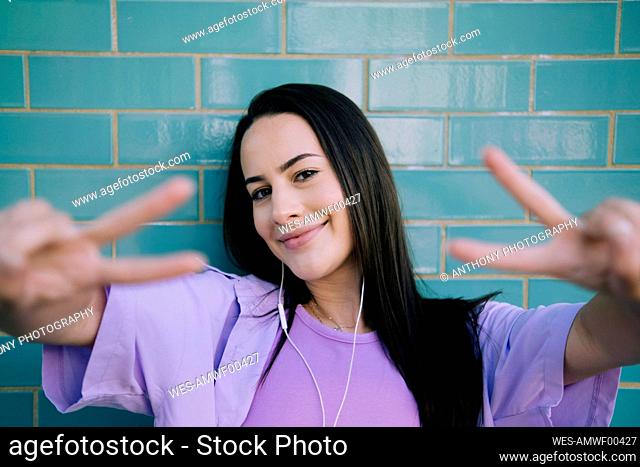 Smiling young woman gesturing peace sign in front of wall