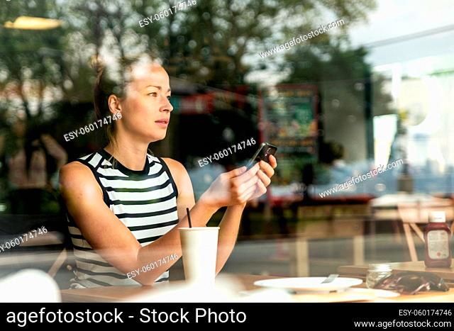 Thoughtful casual caucasian woman holding mobile phone while looking through the coffee shop window during coffee break. Street reflections in the window glass