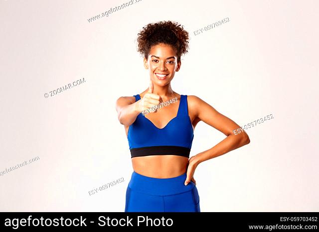 Healthy and fit african-american sportswoman, showing thumbs-up in approval, recommend gym or workout app, standing against white background