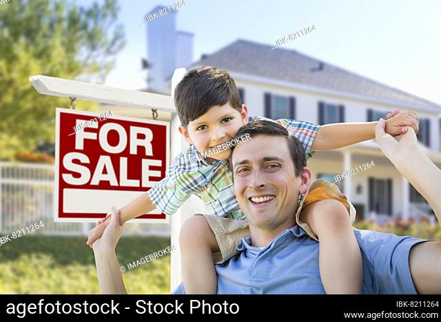 mixed-race father and son piggyback in front house and for sale real estate sign
