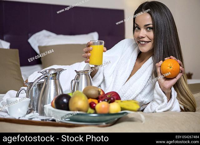 Young beautiful woman in hotel morning gown lying on bed with juice and orange with plate full of fresh fruits laid near