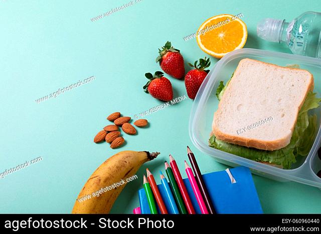Sandwich in lunch box by fruits, almonds and color pencils with books on green background