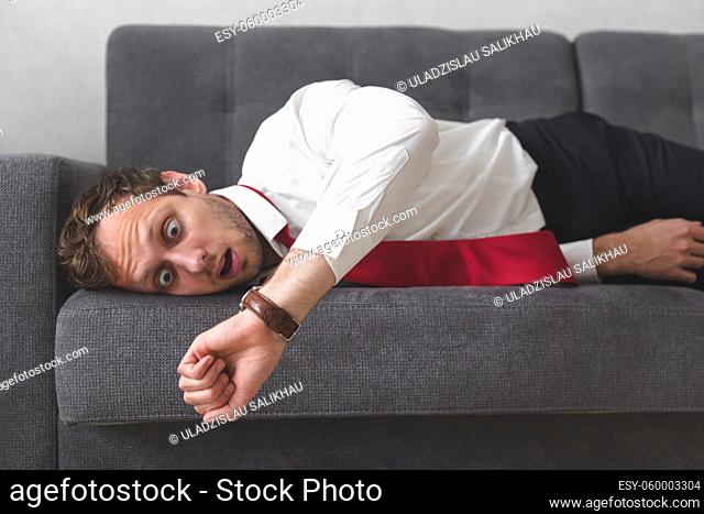 Young office worker overslept and late, looking at watch, lying in office clothing on sofa at home
