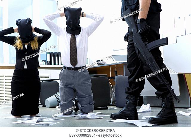 Photo of kneeling businesspeople wearing black sacks on their heads with burglar standing near by