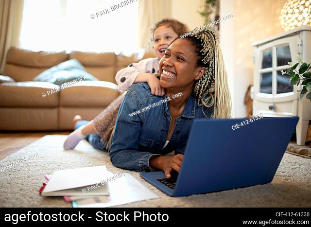 Daughter laying on happy mother working at laptop on floor at home