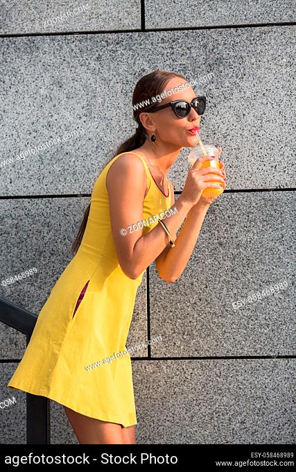 Profile of fashion girl in sunglasses near brick wall. Brown-haired woman tasting orange cockrail. Fashionable model posing in little yellow dress