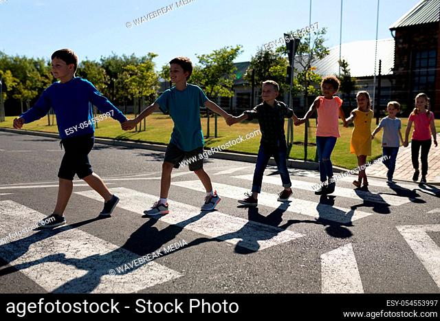Front view of a diverse group of elementary school pupils crossing an empty road together, half way across a pedestrian crossing, holding hands on a sunny day