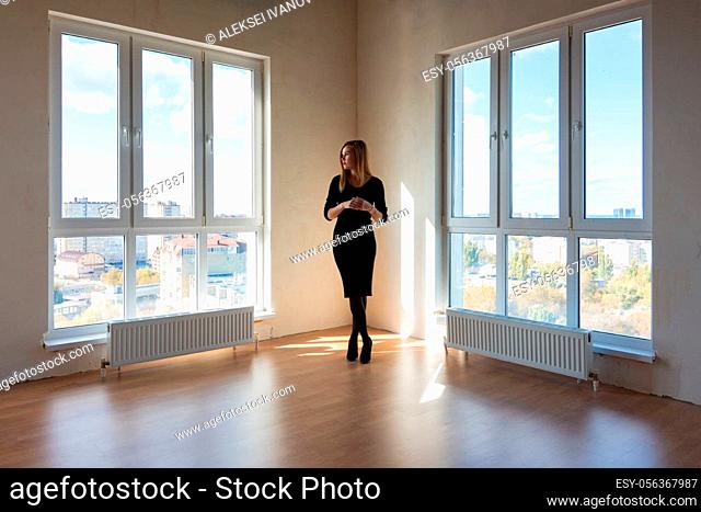 A girl in a black dress stands between two large windows in a spacious empty apartment