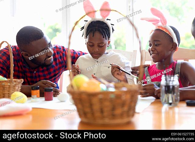 African american father with son and daughter wearing bunny ears painting easter eggs