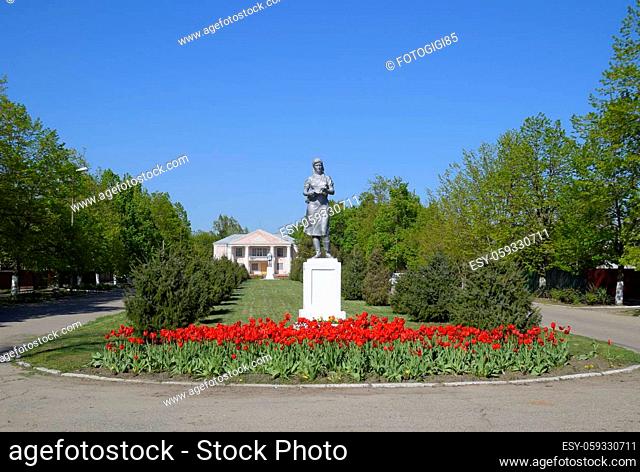 Statue of a collective farmer on a pedestal. The legacy of the Soviet era. A flower bed with tulips and young trees in the village of Oktyabrsky