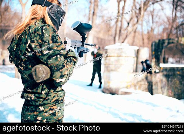 Female paintball player shooting at the enemy, back view, winter forest battle. Extreme sport game, players in protection masks and camouflage lies on the...