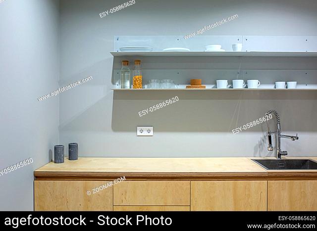 Wooden lockers with a tabletop with a sink in the kitchen in a loft style. Over the tabletop there are shelves with cups, glasses, bottles and plates