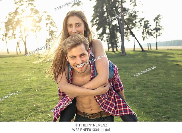 Portrait of happy young man carrying girlfriend piggyback on meadow