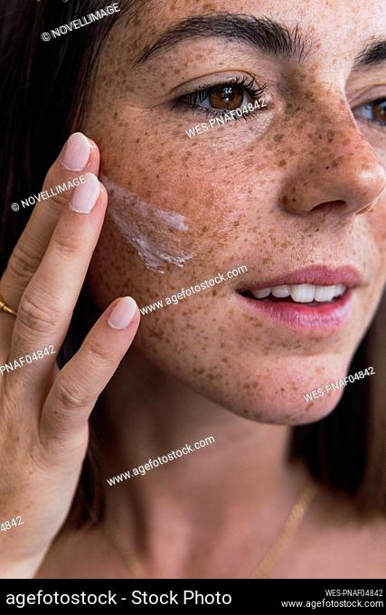Young woman with freckle face applying moisturizer on face